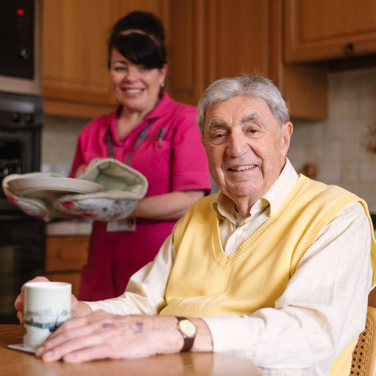 Lady with a care assistant job smiling whilst providing care for an elderly male client
